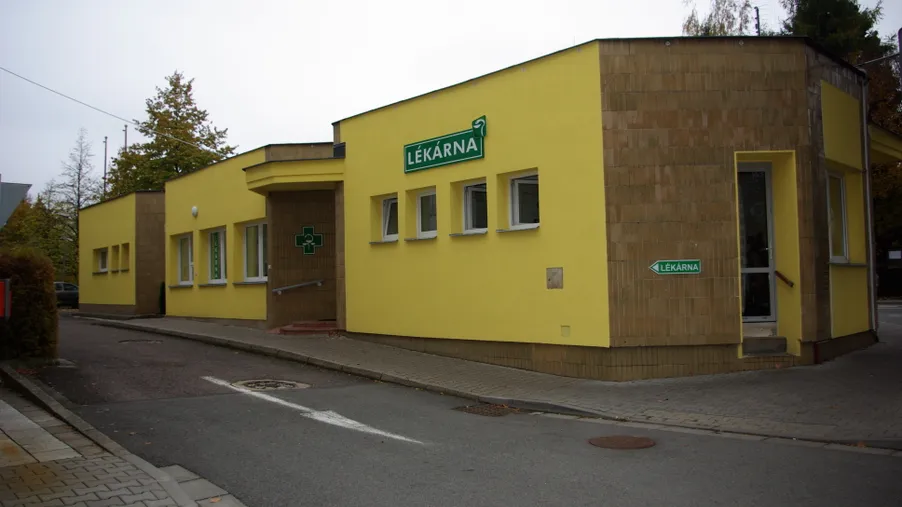 Exteriér jednopodlažní žluté lékárny umístěné na rohu ulice s nápisem „LÉKÁRNA“ na fasádě.